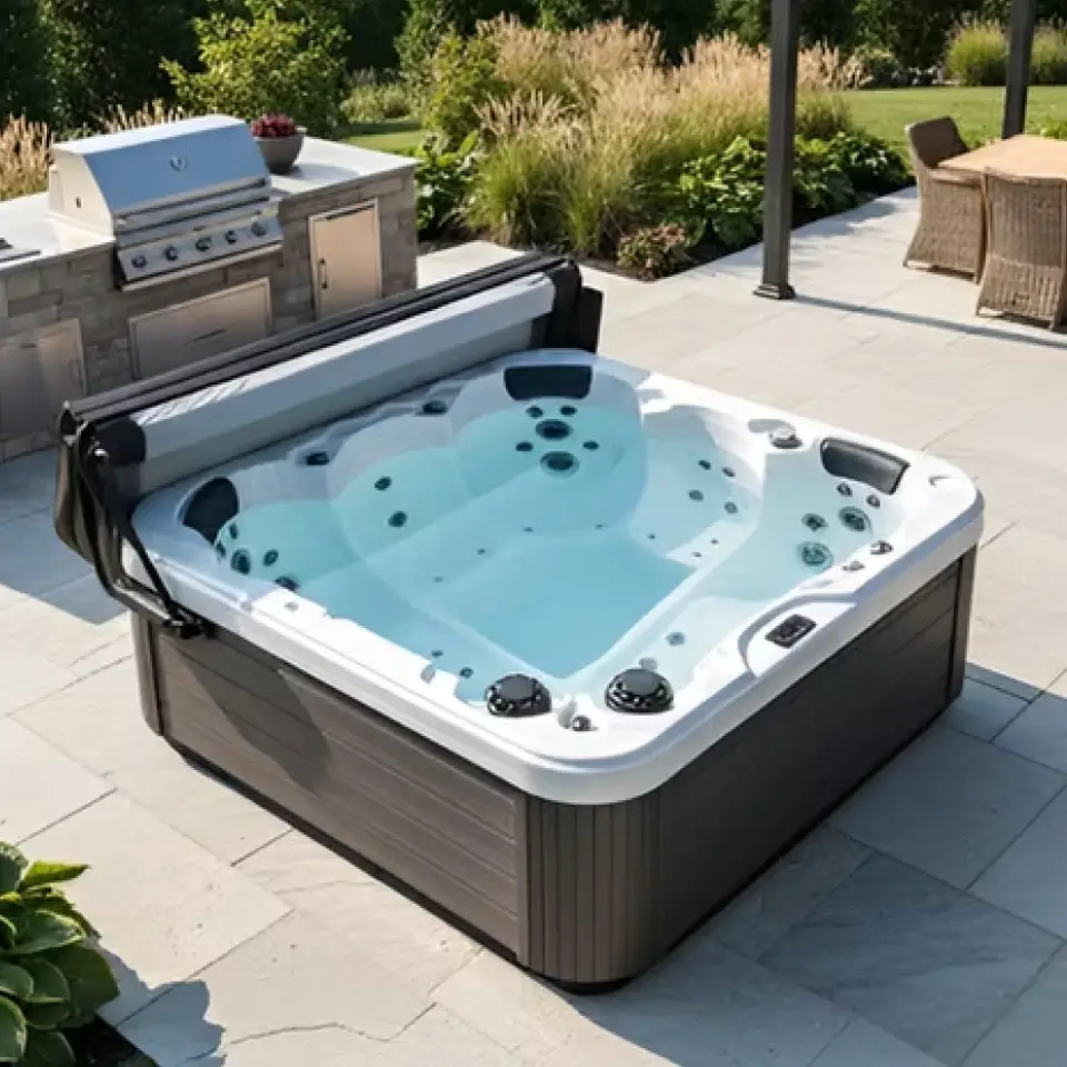 A spa with wooden style texture on the sides and white marble texture for the top and interior sits on a tiled patio in an outdoor area with greenery, seating and a grill around. The spa has a fully-opened roll-away cover.